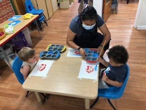 Person in mask helping two children paint at a table paint