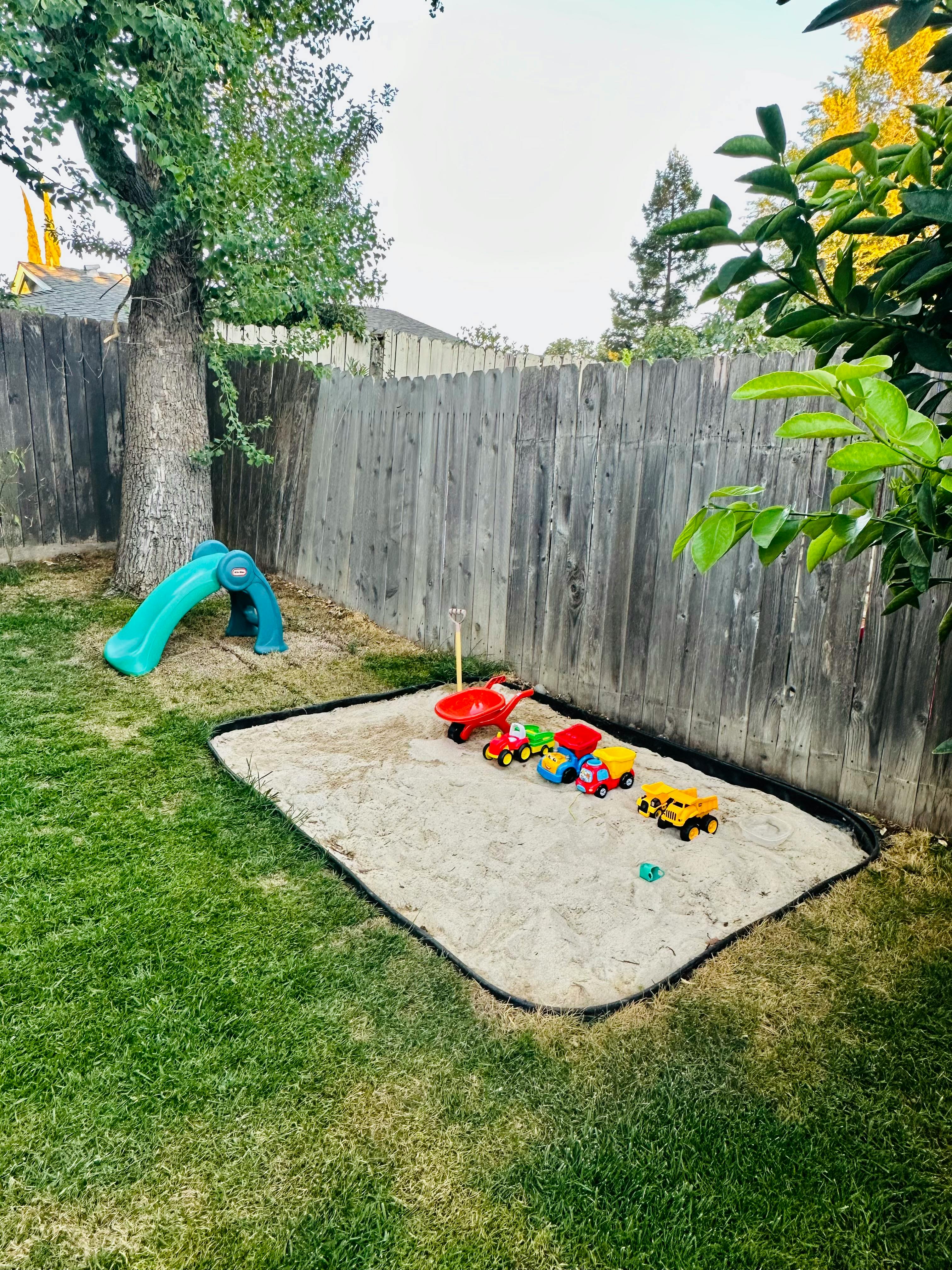 Sandbox with toy trucks and a red wheelbarrow next to a small slide