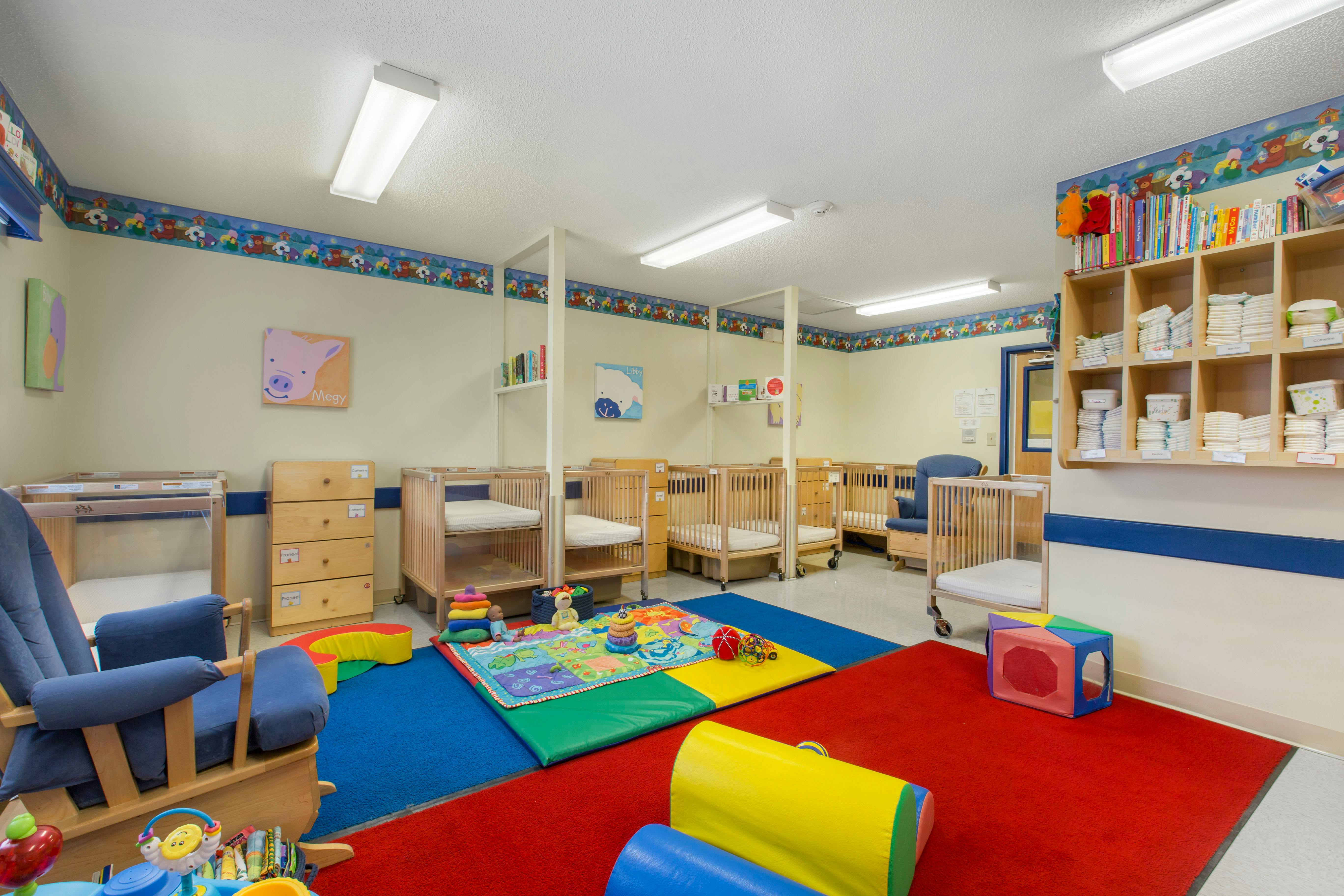 A childcare room with multiple cribs, a blue rocking chair, and colorful play mats and toys