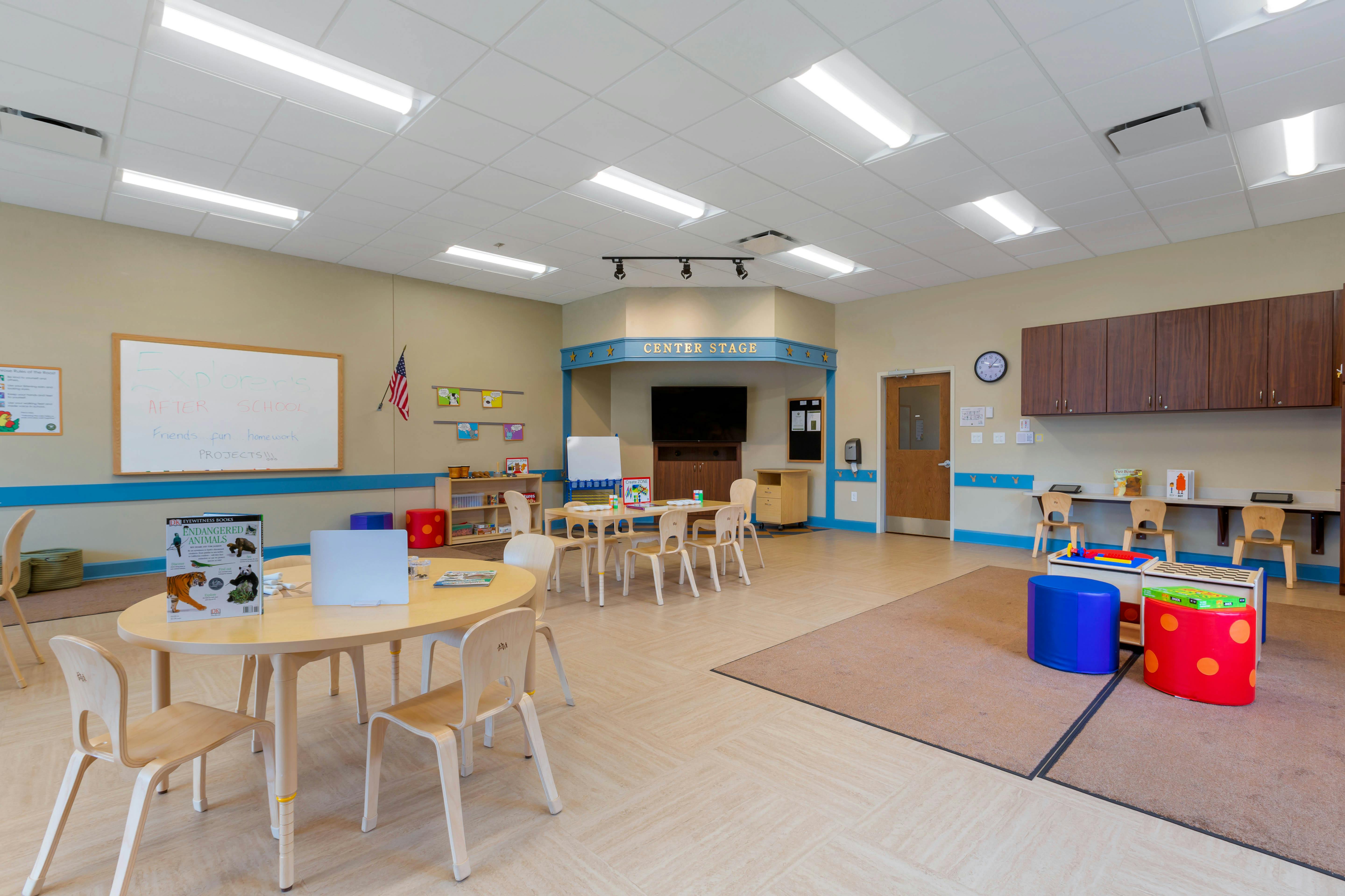An empty after-school classroom with small tables and chairs, a whiteboard, and a small stage