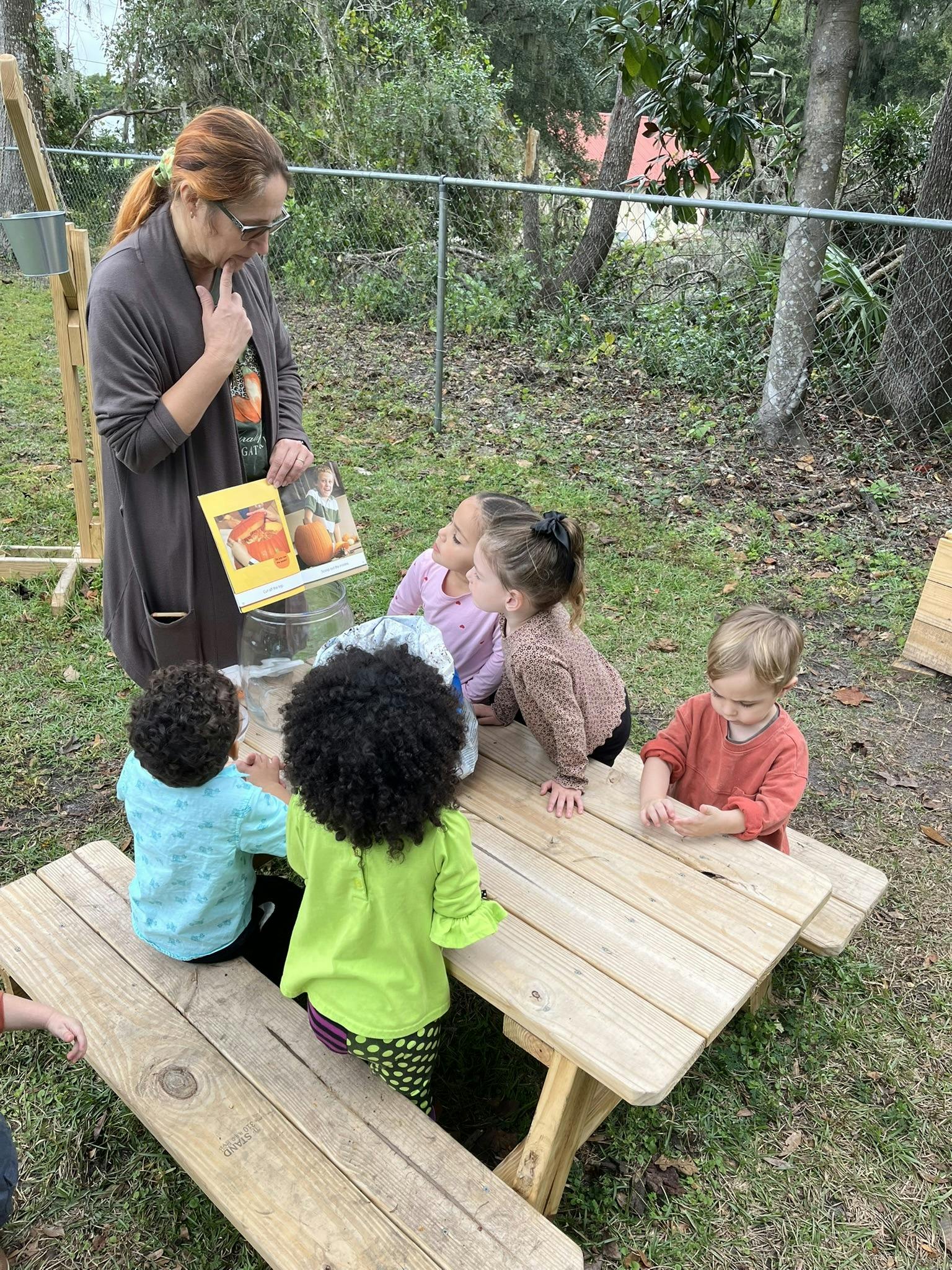 Person reading a book to children sitting at a picnic table outdoors