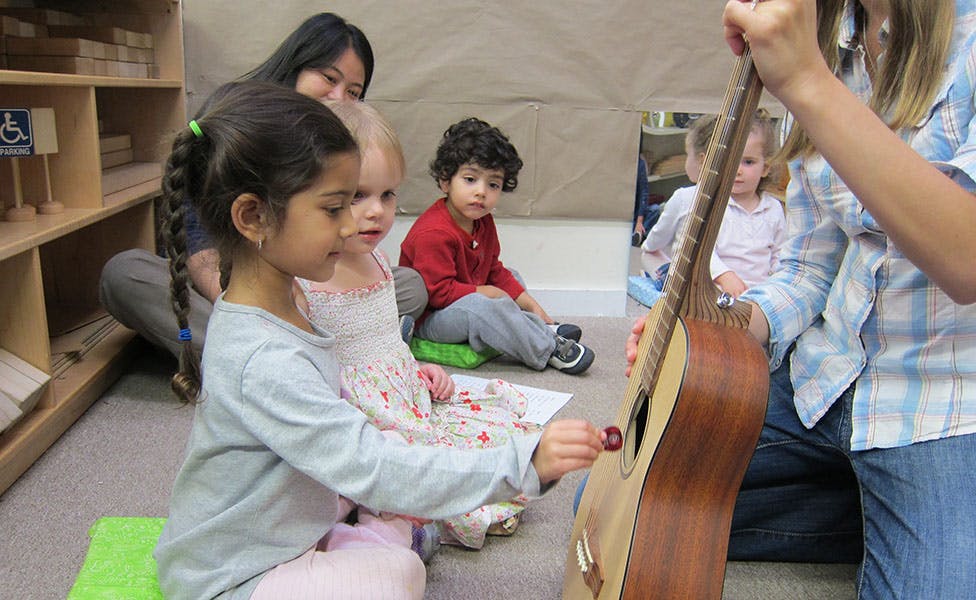 Child touching a guitar held by a person, with other children and adults nearby