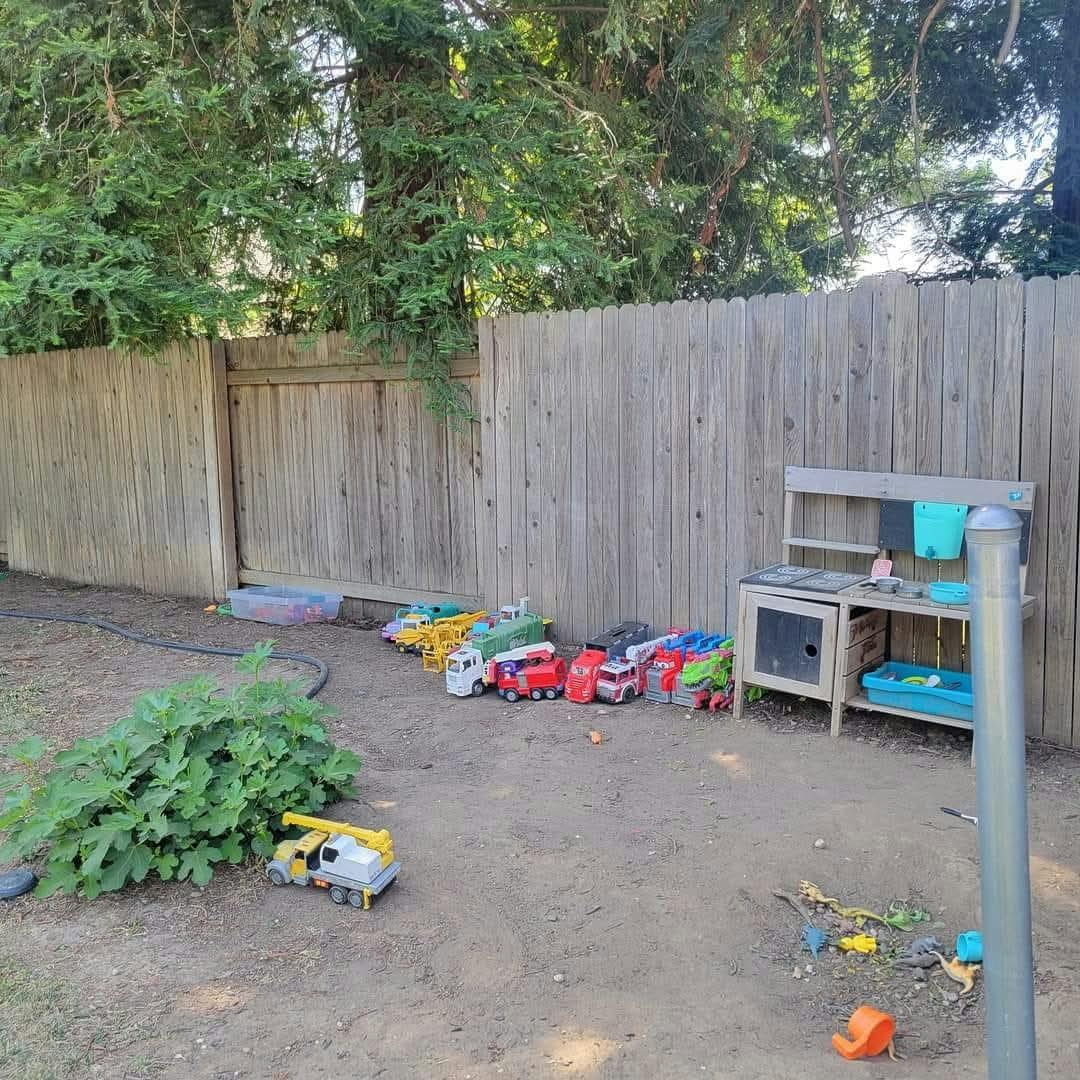 Toy trucks and a play kitchen in a dirt yard next to a wooden fence