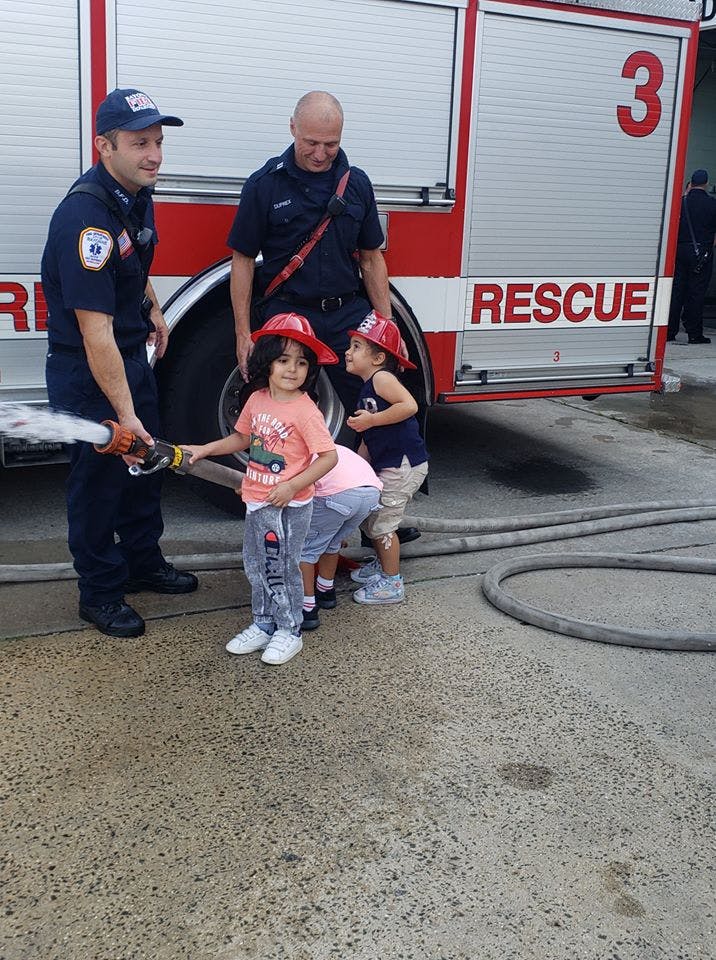 Two adults in uniform with three children in red hats holding a fire hose