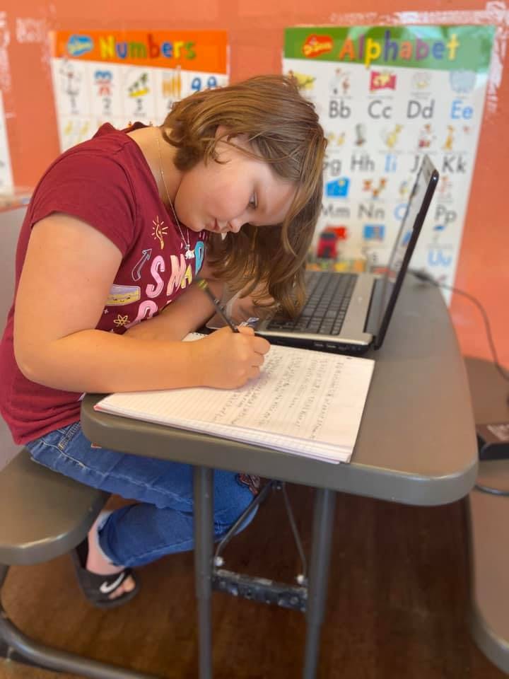 Child writing on paper at a desk with a laptop