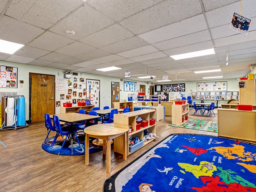 Classroom with tables, blue chairs, educational rugs, and shelves with wall art