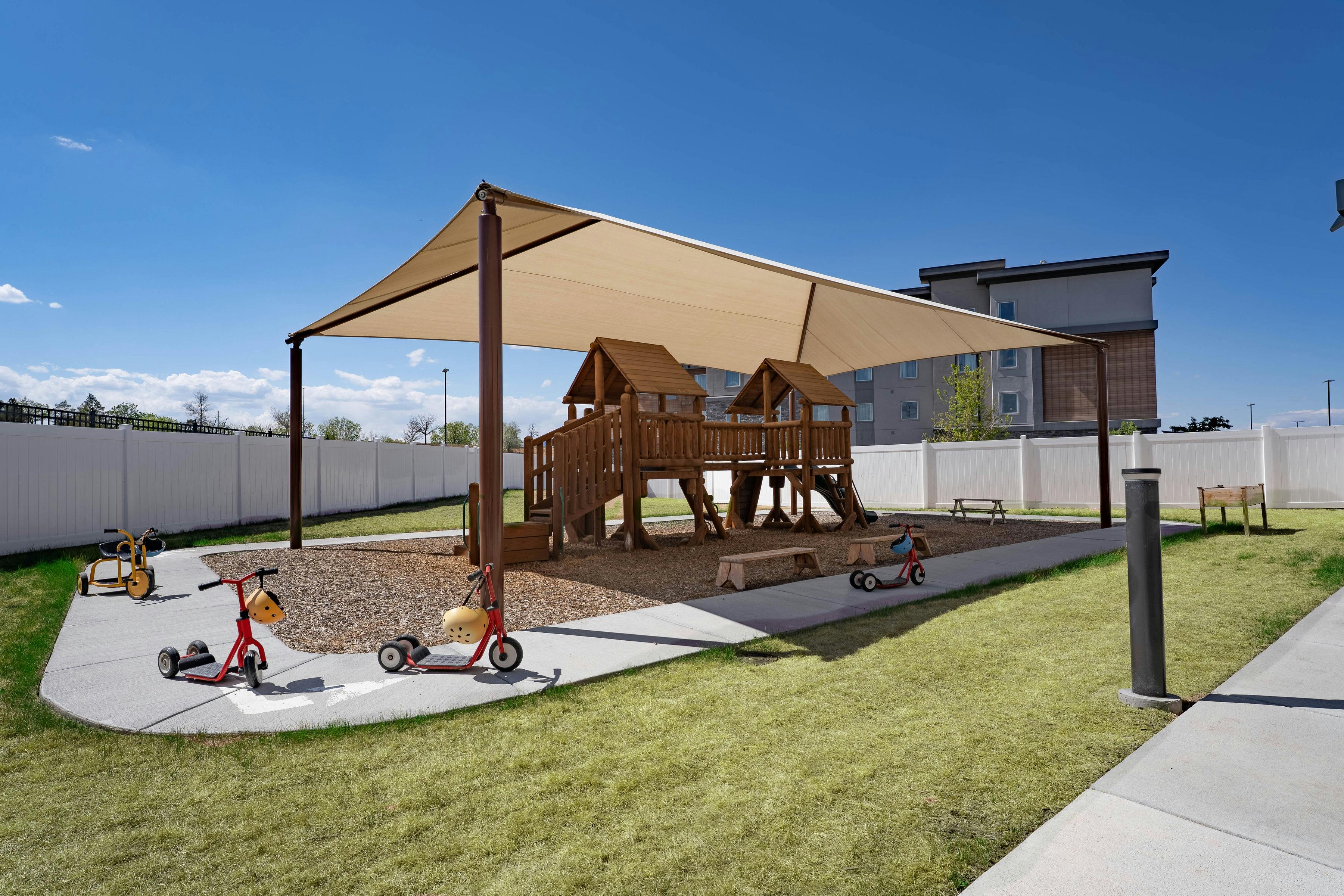 Playground with wooden playset under a shaded canopy and tricycles on a path