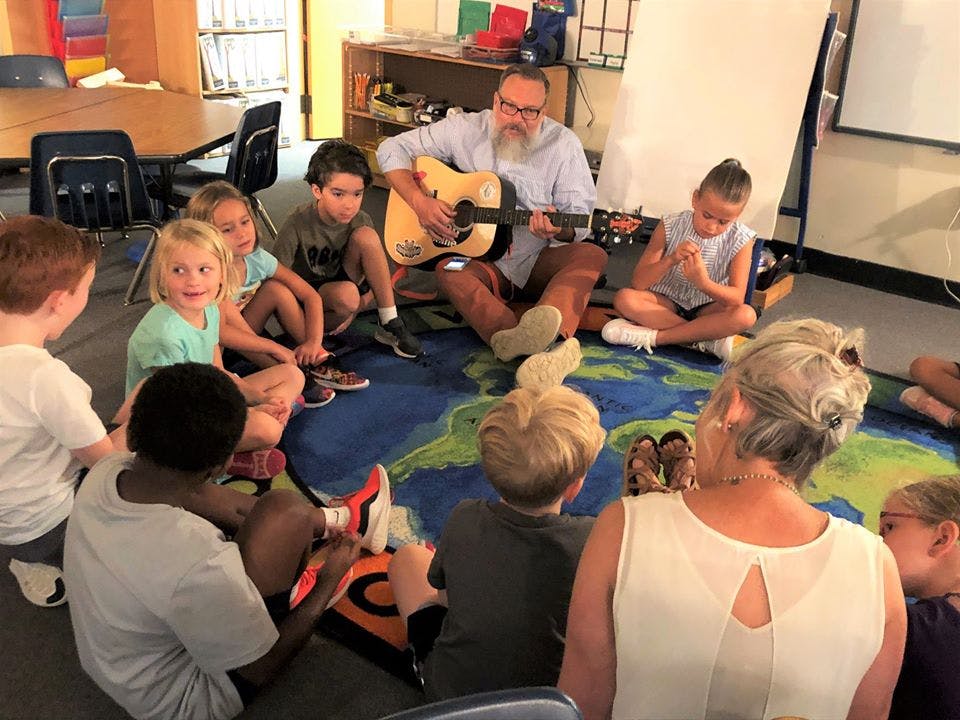 Person playing guitar for a group of children and adults sitting on a rug