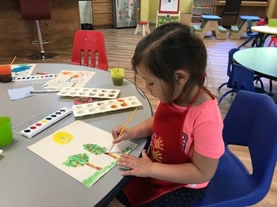 Child painting a picture of trees and a sun at a table