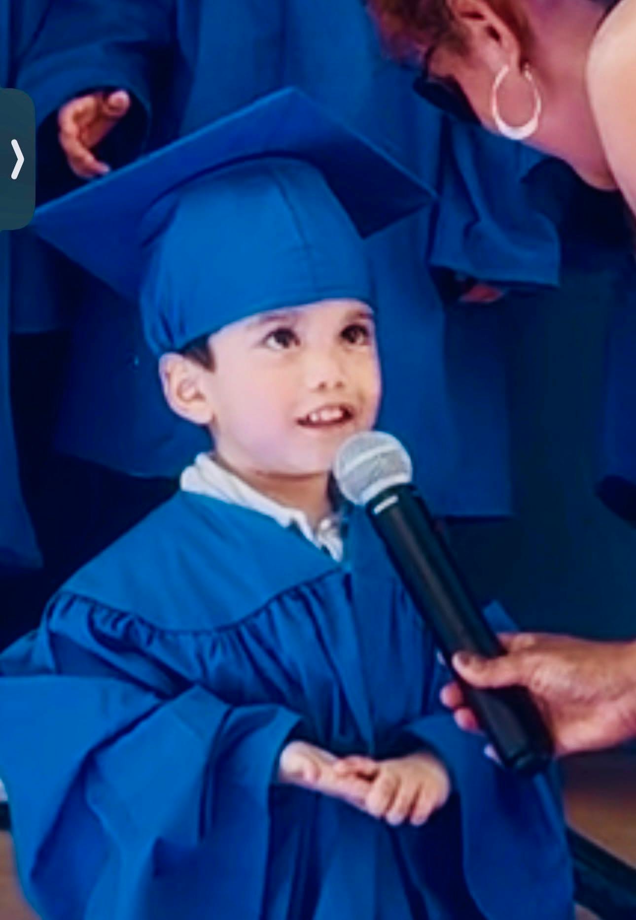 Child in blue graduation cap and gown speaking into a microphone