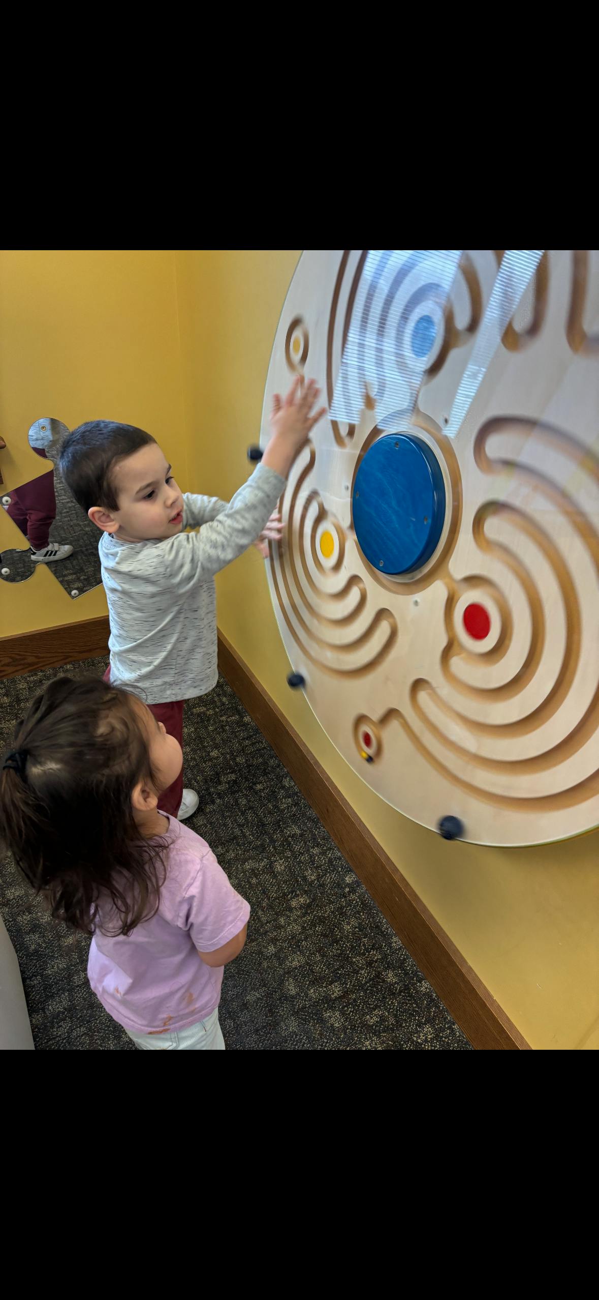 Two children playing with a circular wall-mounted marble maze game