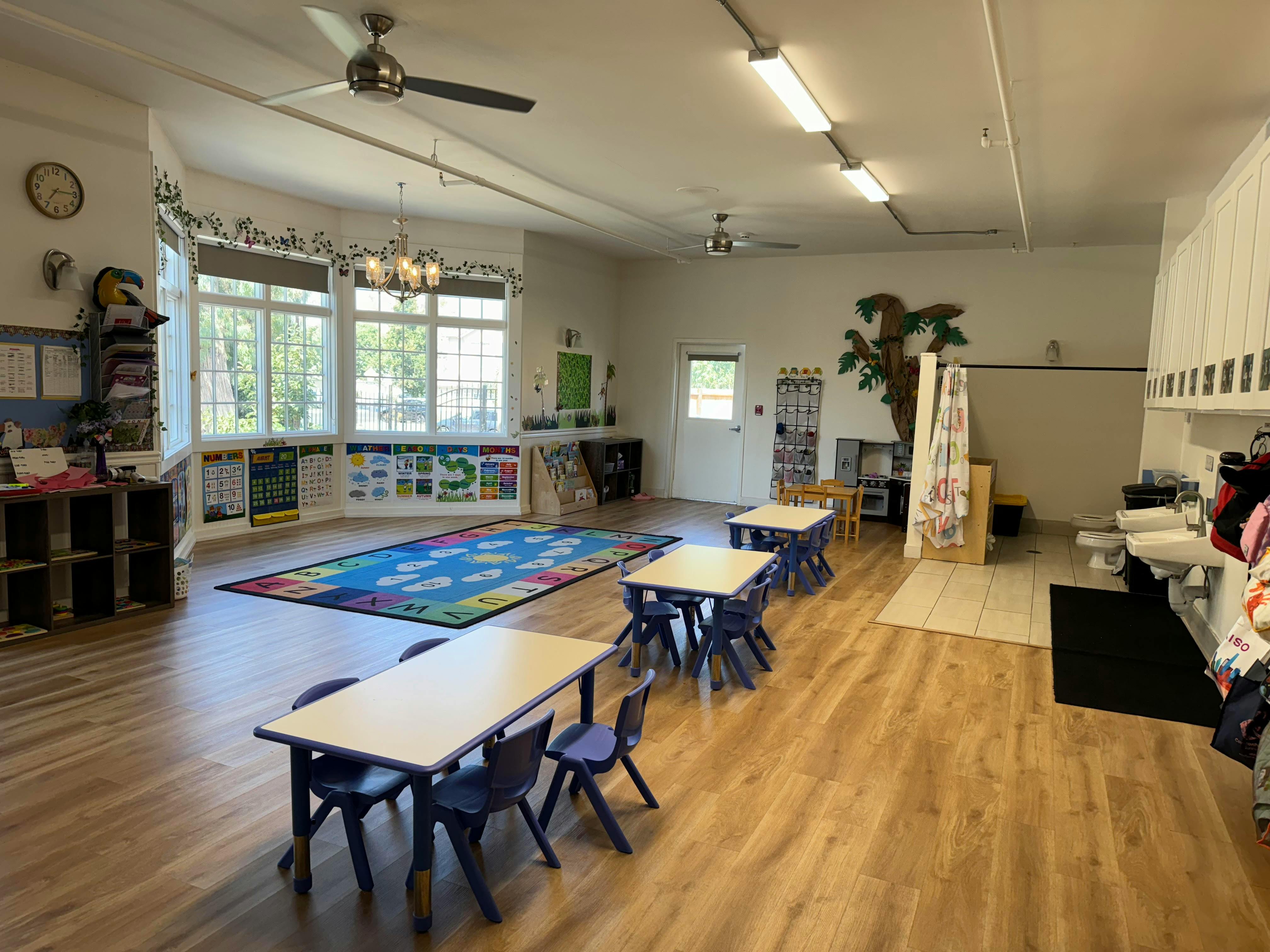 Empty classroom with small tables and chairs, a colorful rug, and large windows