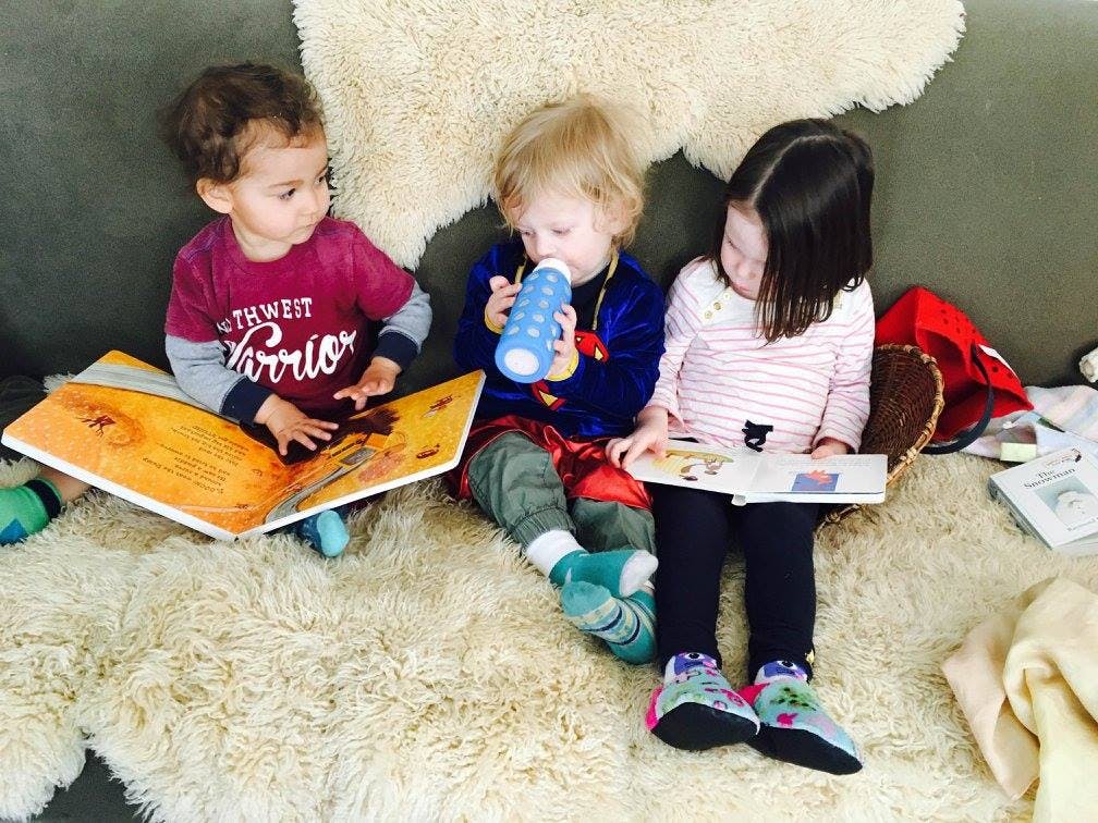 Three children sitting on a couch, one looking at a book, one drinking, one reading