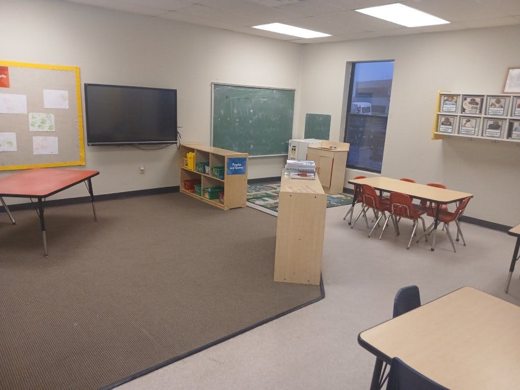 Empty classroom with tables, chairs, a blackboard, and a wall-mounted television