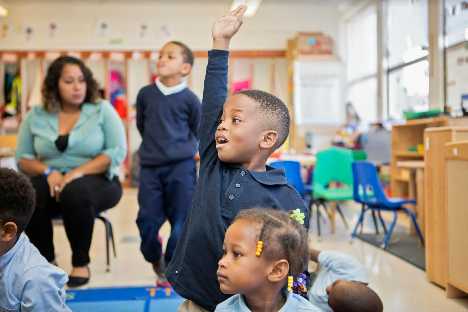 Child raising hand in a classroom with other children and an adult
