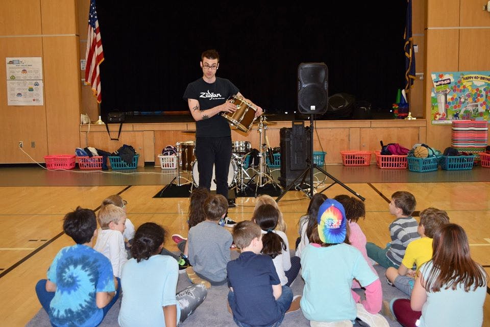 Person on stage holding a drum, children sitting on floor watching