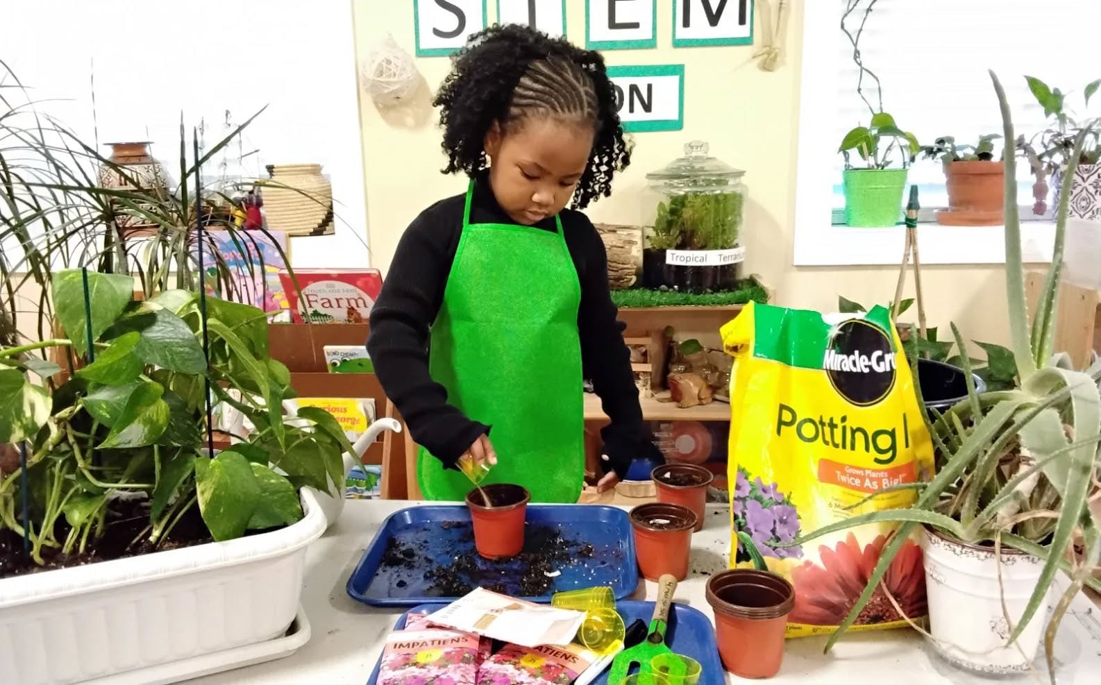 Child in a green apron pouring dirt into a small pot in a classroom