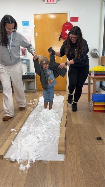 Two people helping a child walk through artificial snow