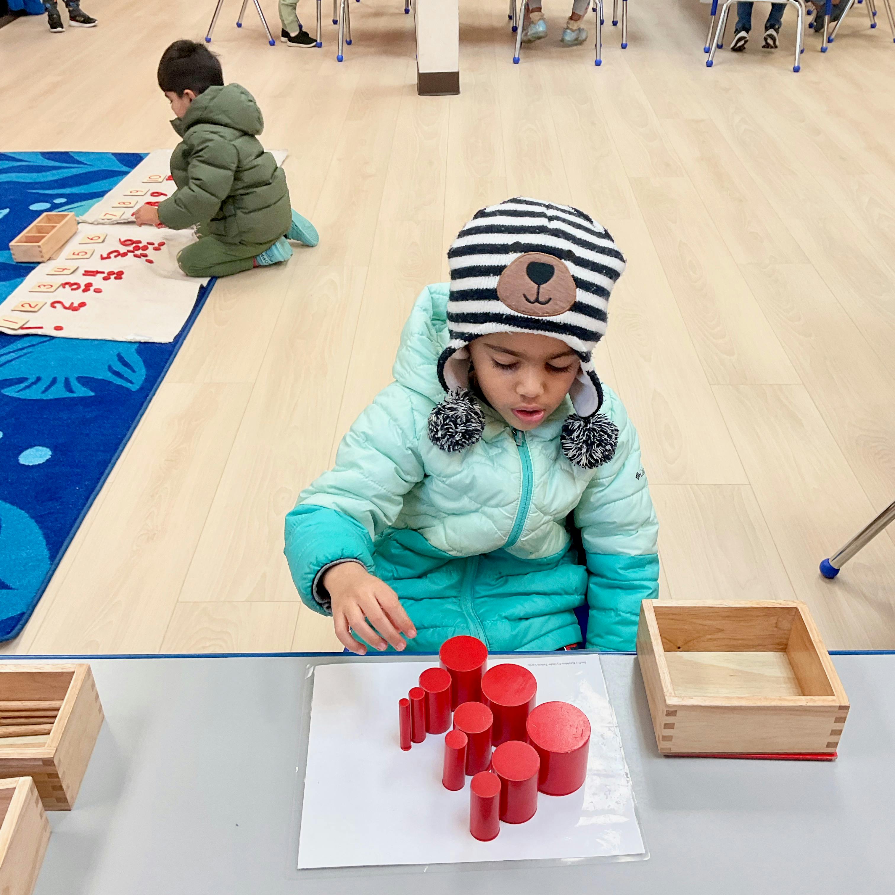 Child in a bear hat playing with red cylindrical blocks insets in a classroom