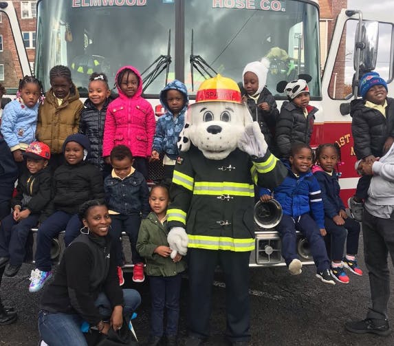 Children and adults posing with a dog mascot in a firefighter uniform in front of a fire truck
