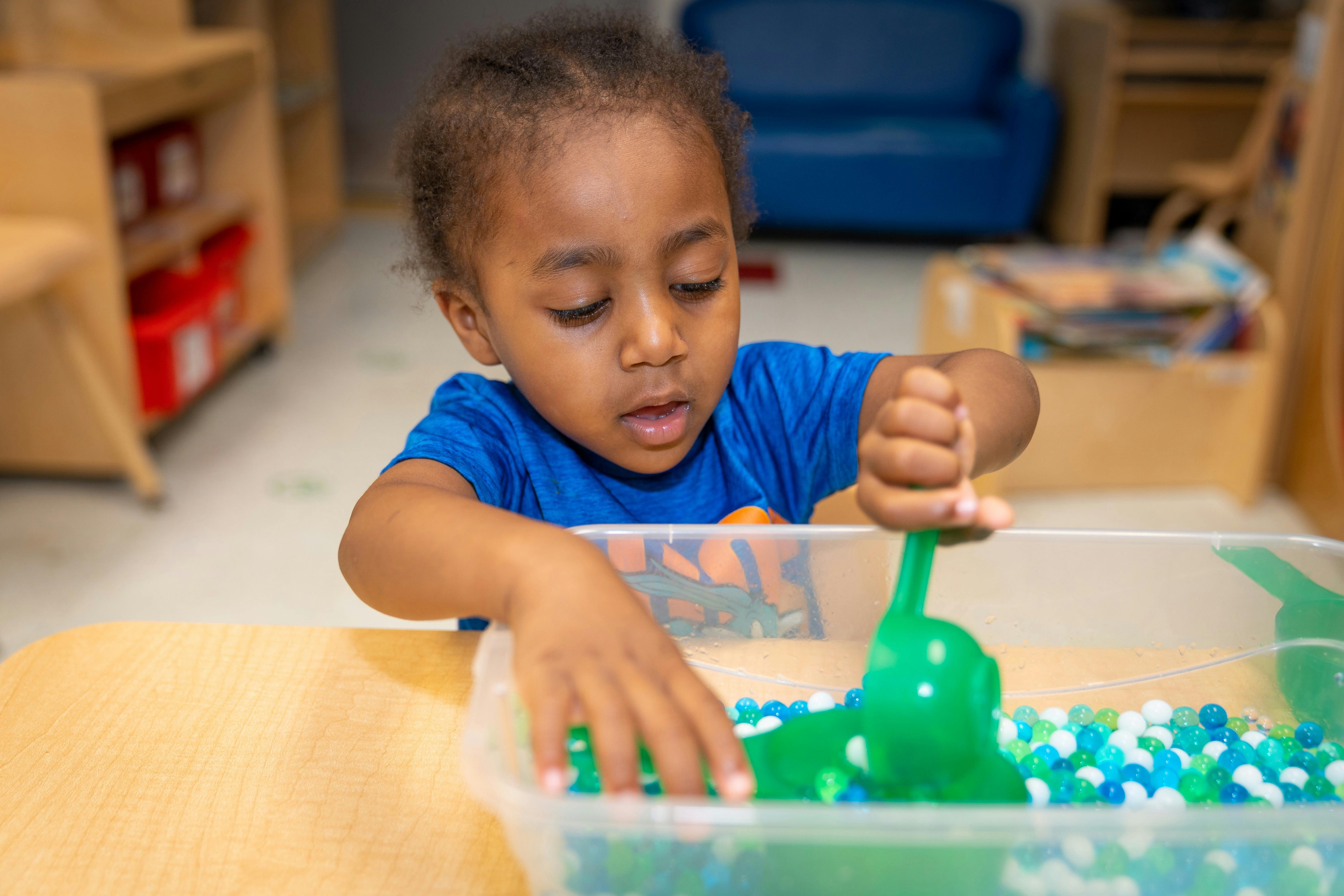 Child playing with a green scoop and colorful beads in a clear bin