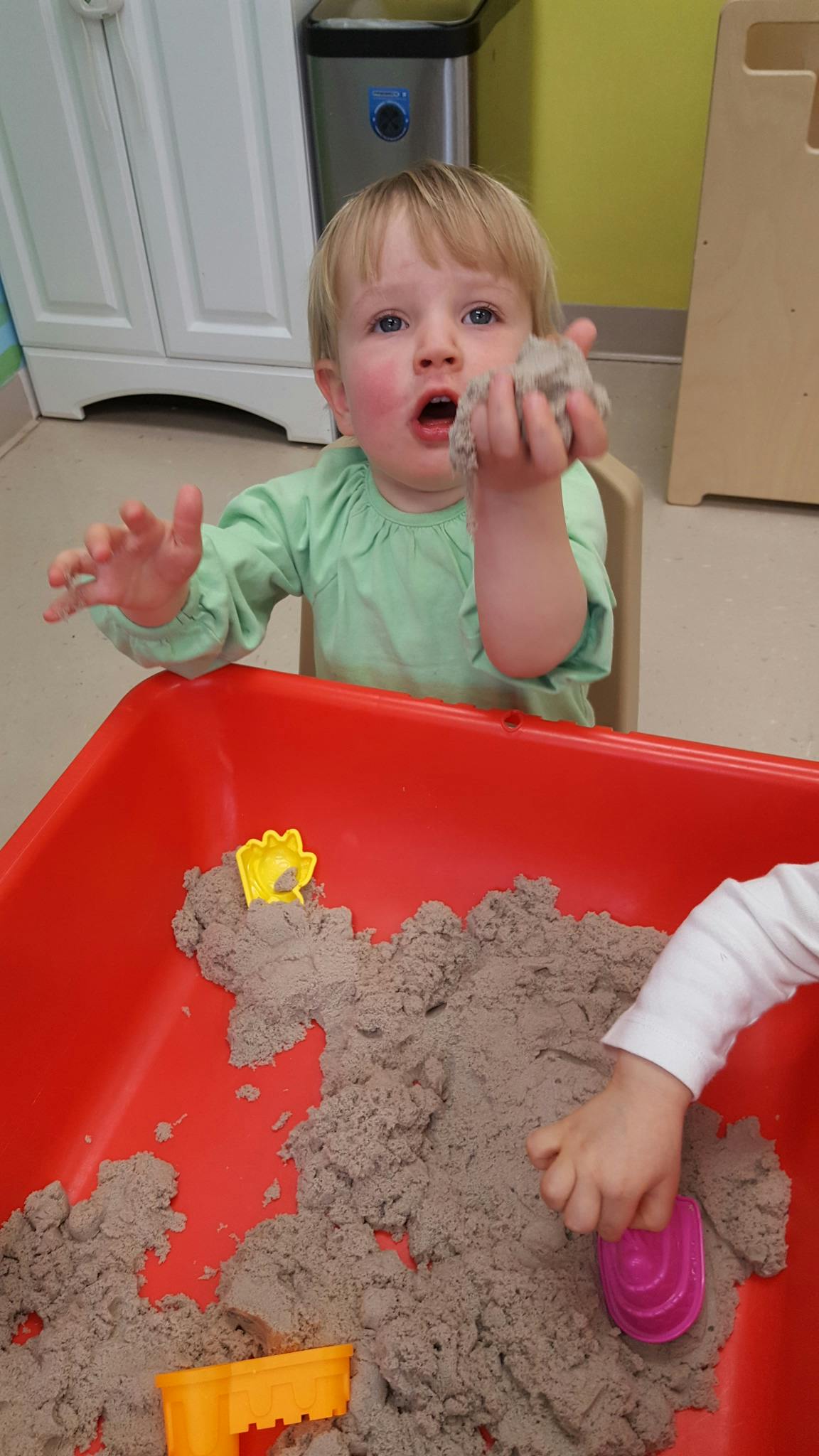 Child holding kinetic sand, another child playing in a red bin of kinetic sand