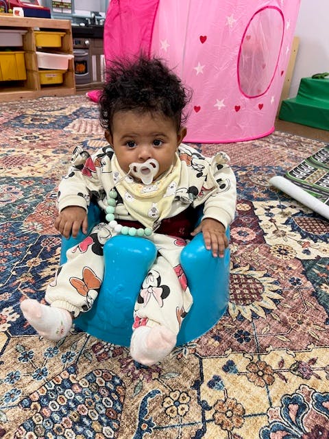Child with pacifier sitting in a blue baby seat on a patterned rug