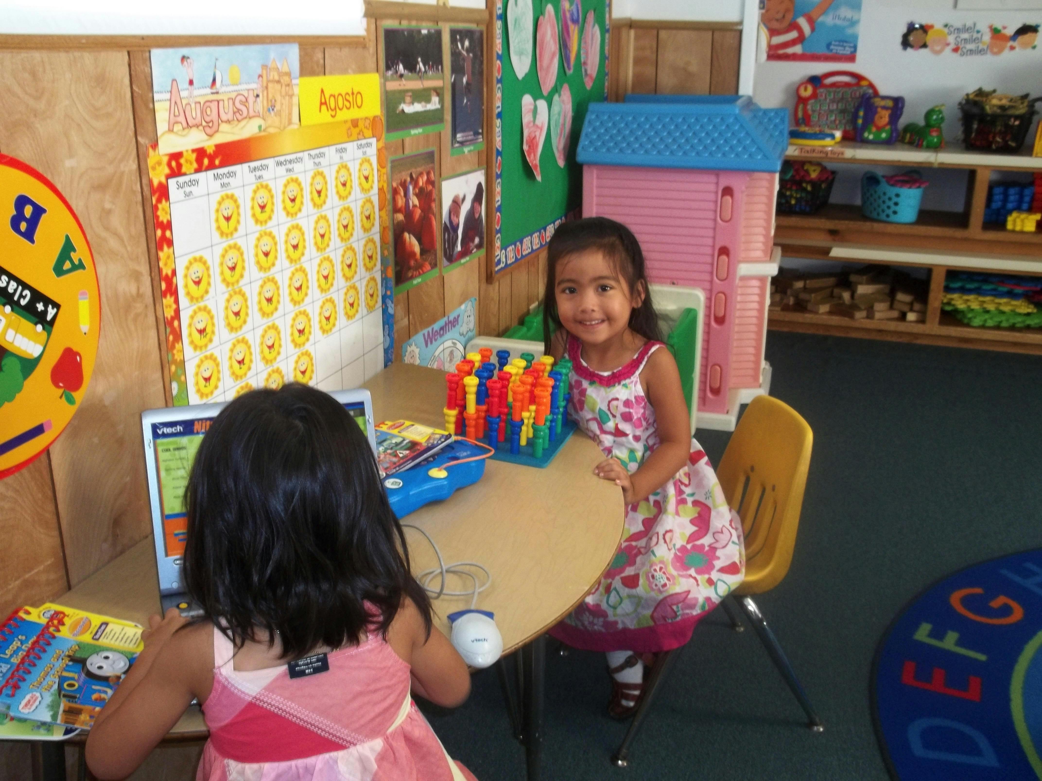 Two children at a table in a classroom, one looking at a laptop
