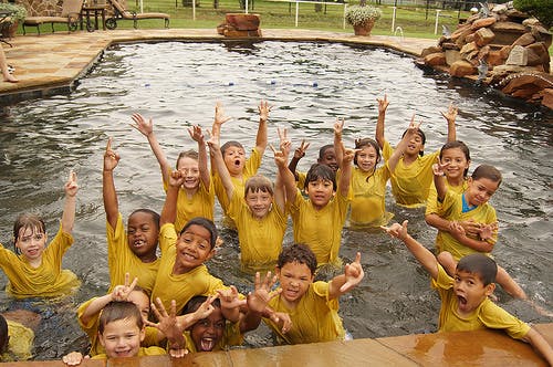 Group of children in yellow shirts in a swimming pool with hands raised