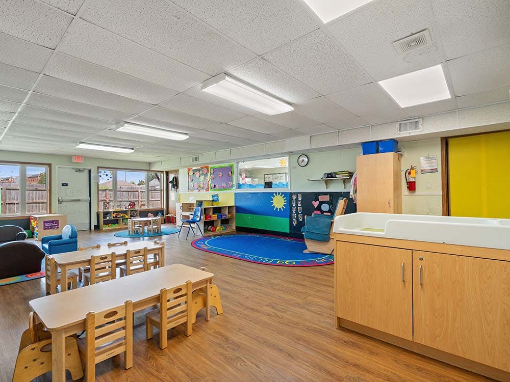 A colorful classroom with tables and chairs for children, a changing station, and play areas