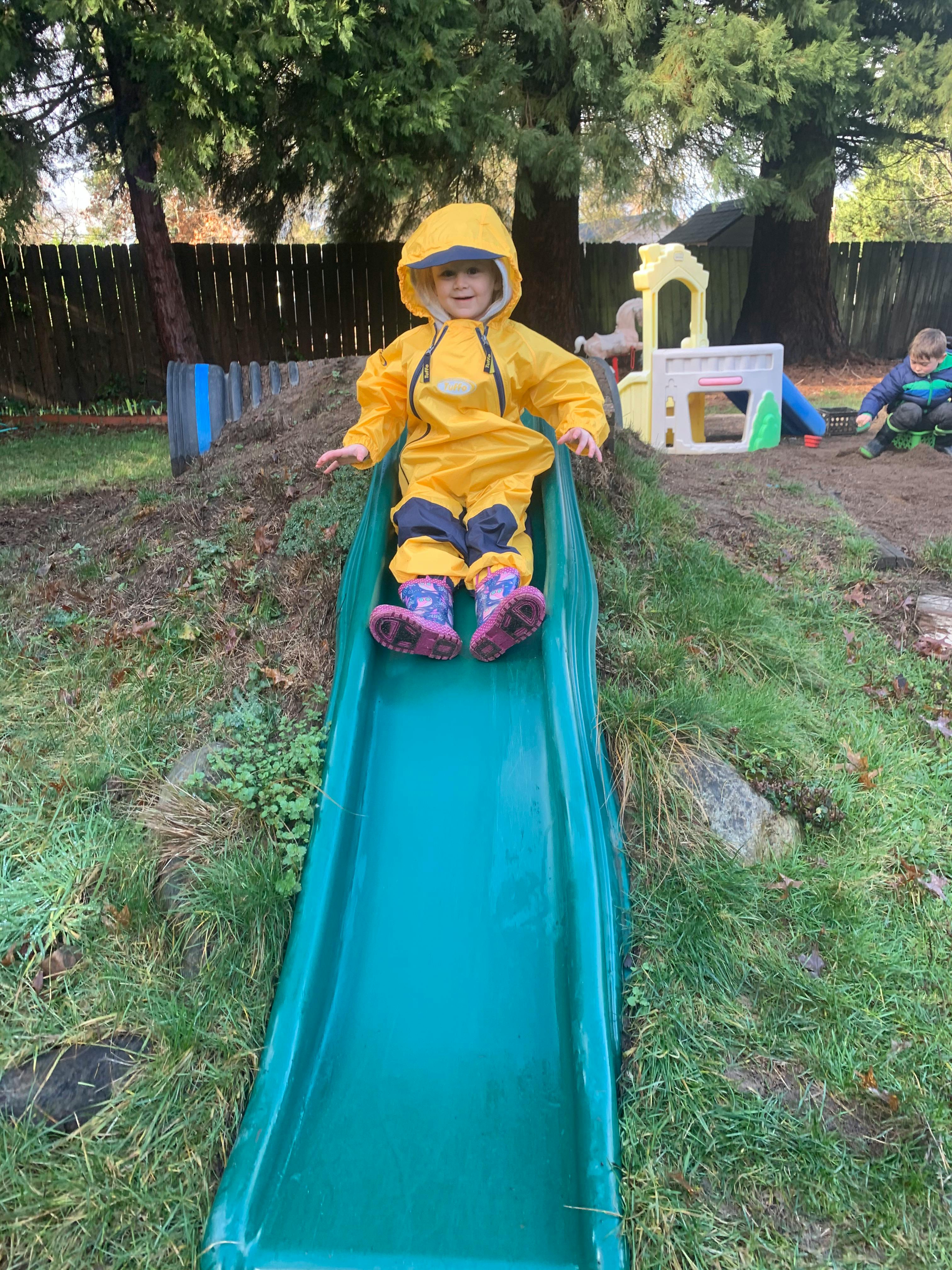 Child in yellow rain suit sitting at the top of a green slide