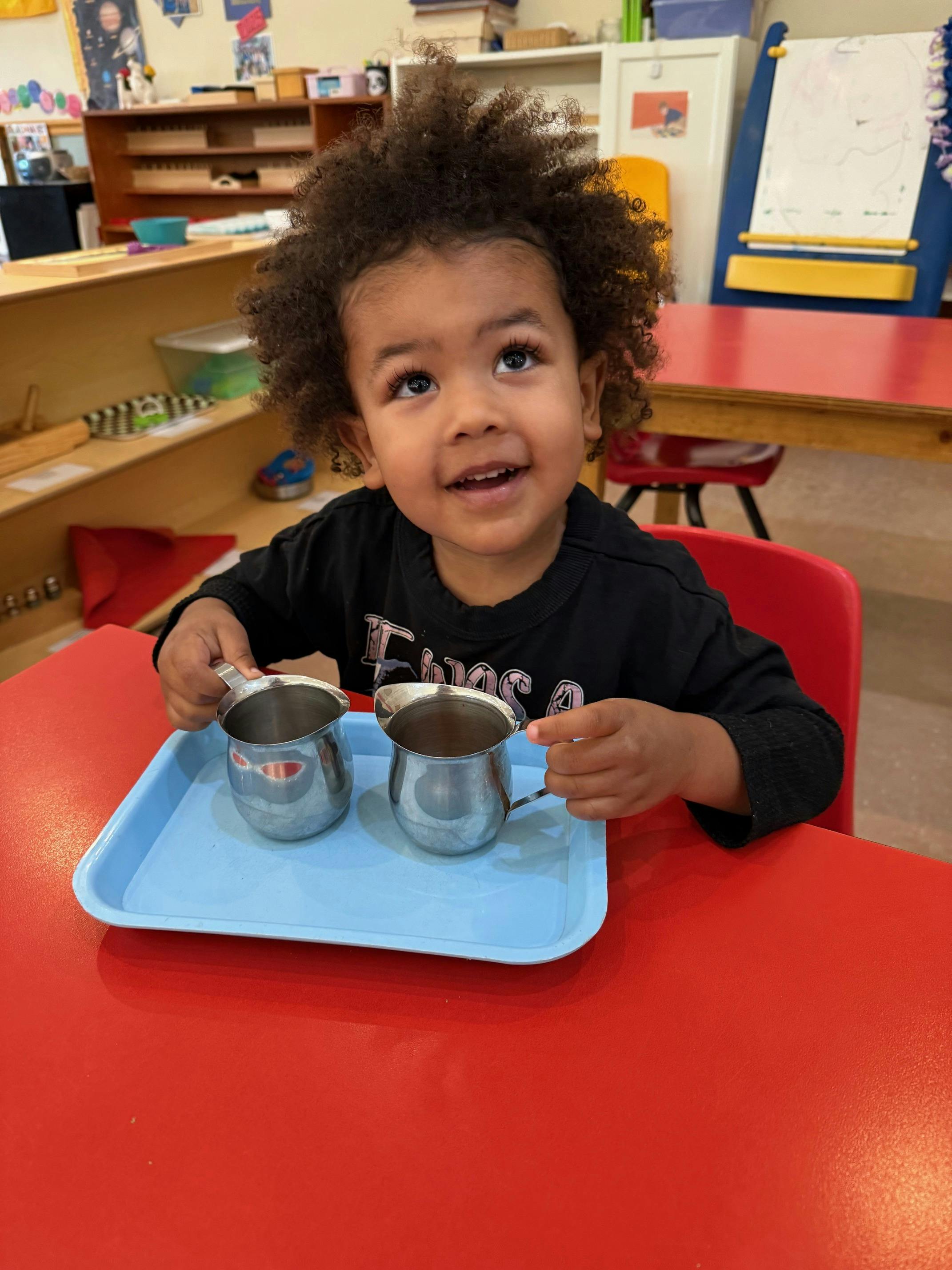 Child sitting at a table with two small metal pitchers on a blue tray