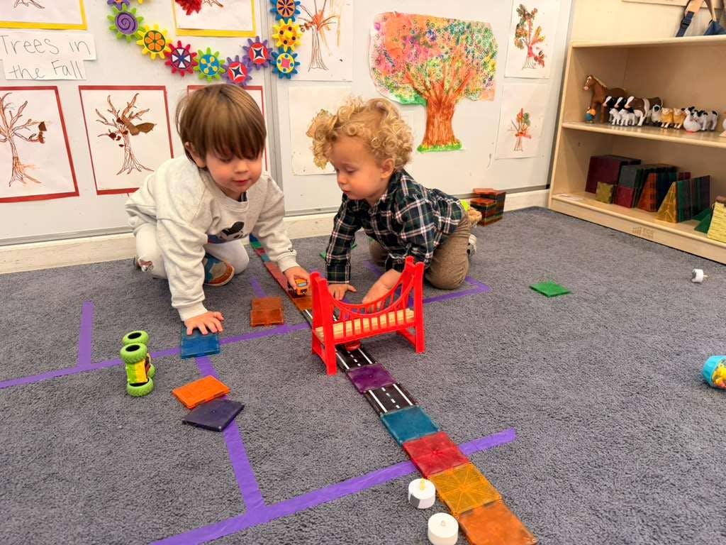 Two children playing with toy cars and magnetic tiles on the floor of a classroom