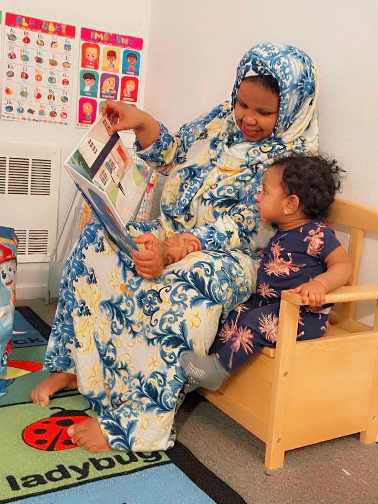 Person in patterned clothing reading a book to a child in a small chair