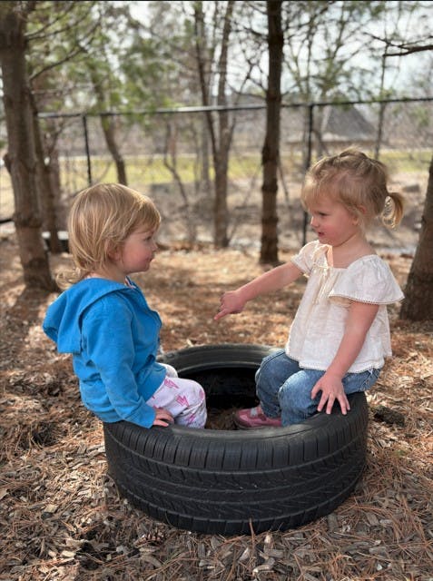 Two children sitting in car tires outdoors, one pointing at the other