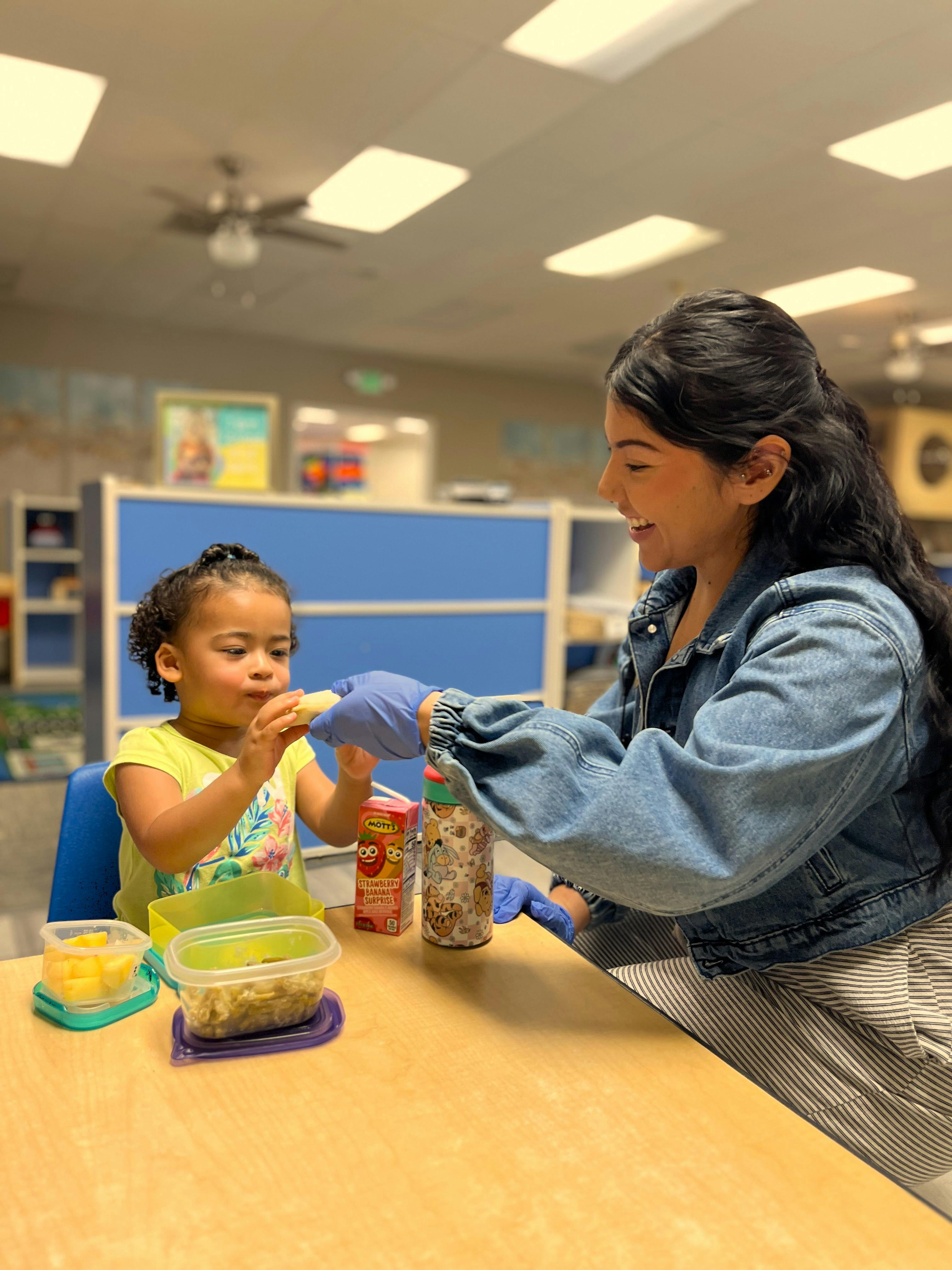 Person feeding a child a snack at a table in a classroom