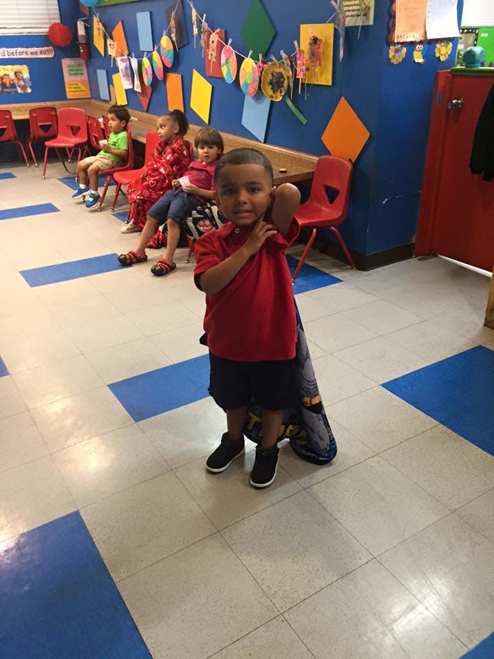 Child standing in a classroom with other children seated in the background