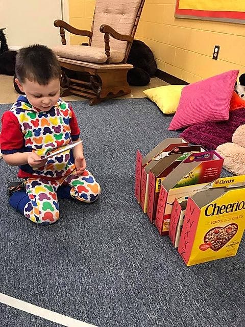 Child kneeling on a rug looking at a small card, next to cereal boxes