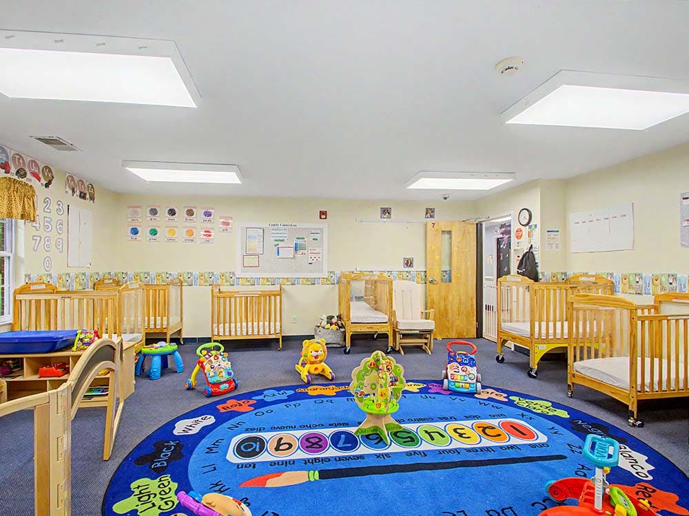 cribs in a brightly lit daycare room with a colorful ABC and 123 rug