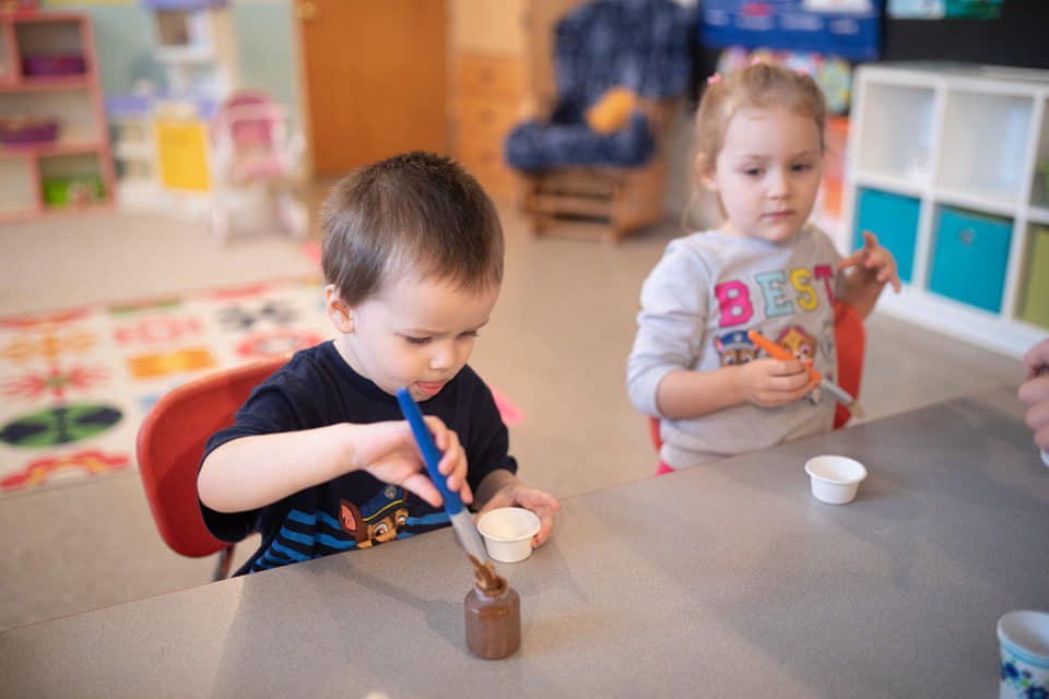 Two children at a table, one painting a small bottle, the other holding a cup
