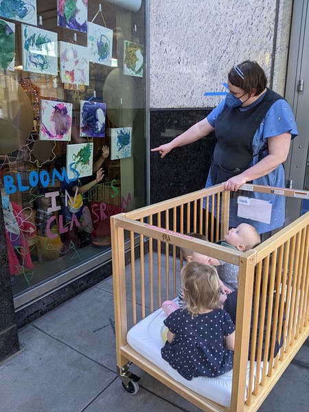 Person pointing at children's artwork displayed in a window with children in a portable crib