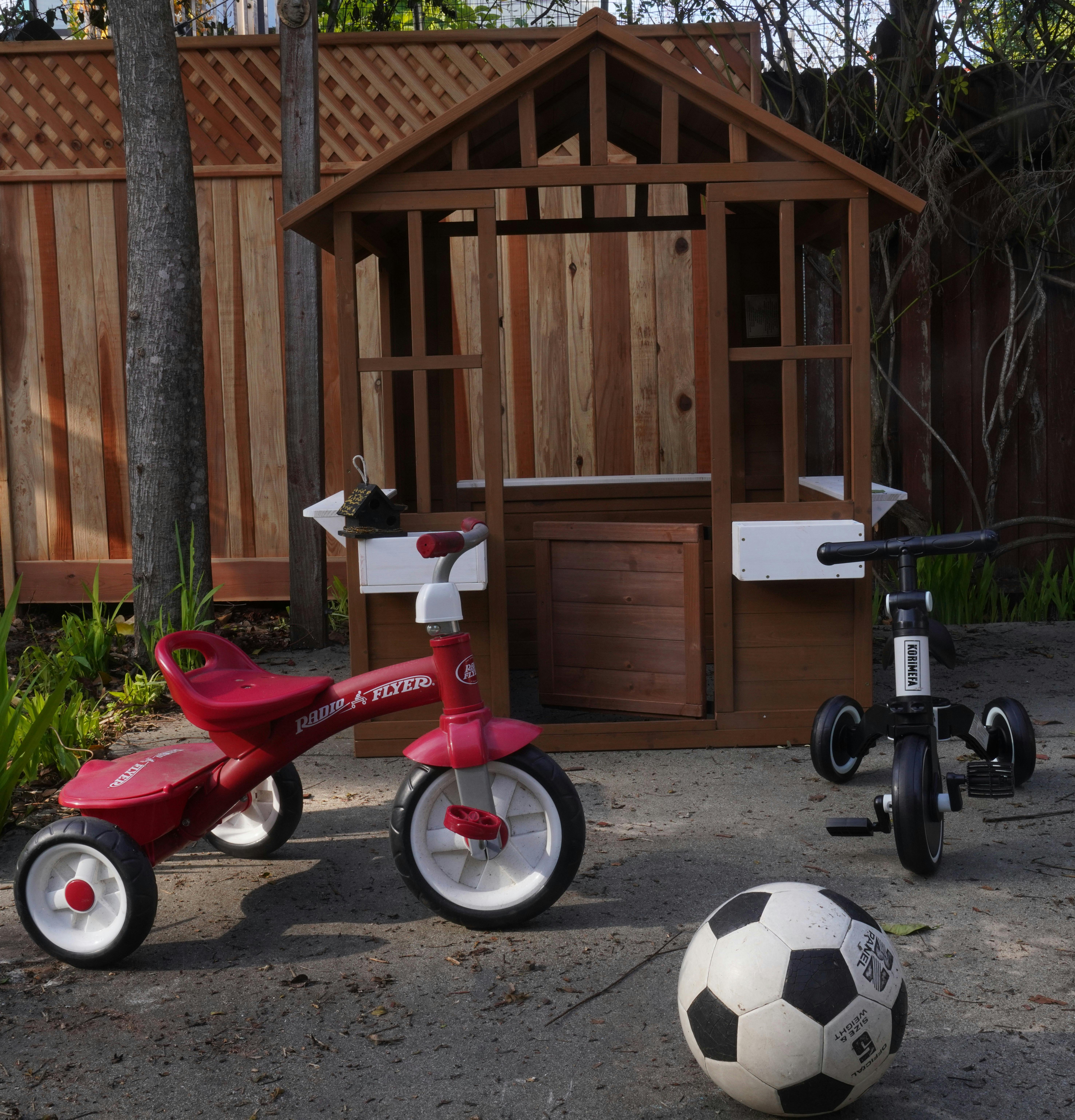 Red tricycle, black scooter, and soccer ball in front of a wooden playhouse