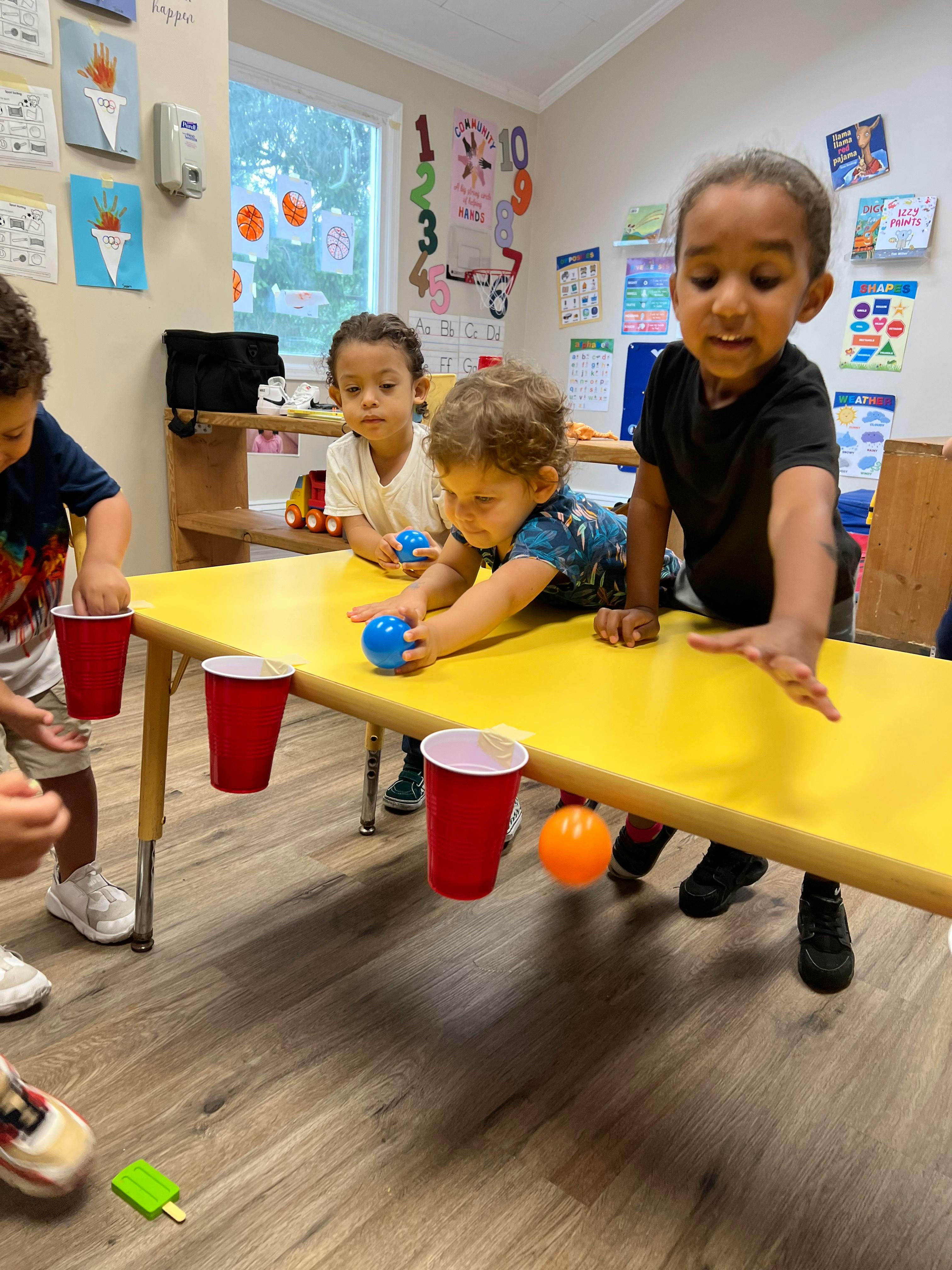 Children playing with balls and red cups on a yellow table in a classroom