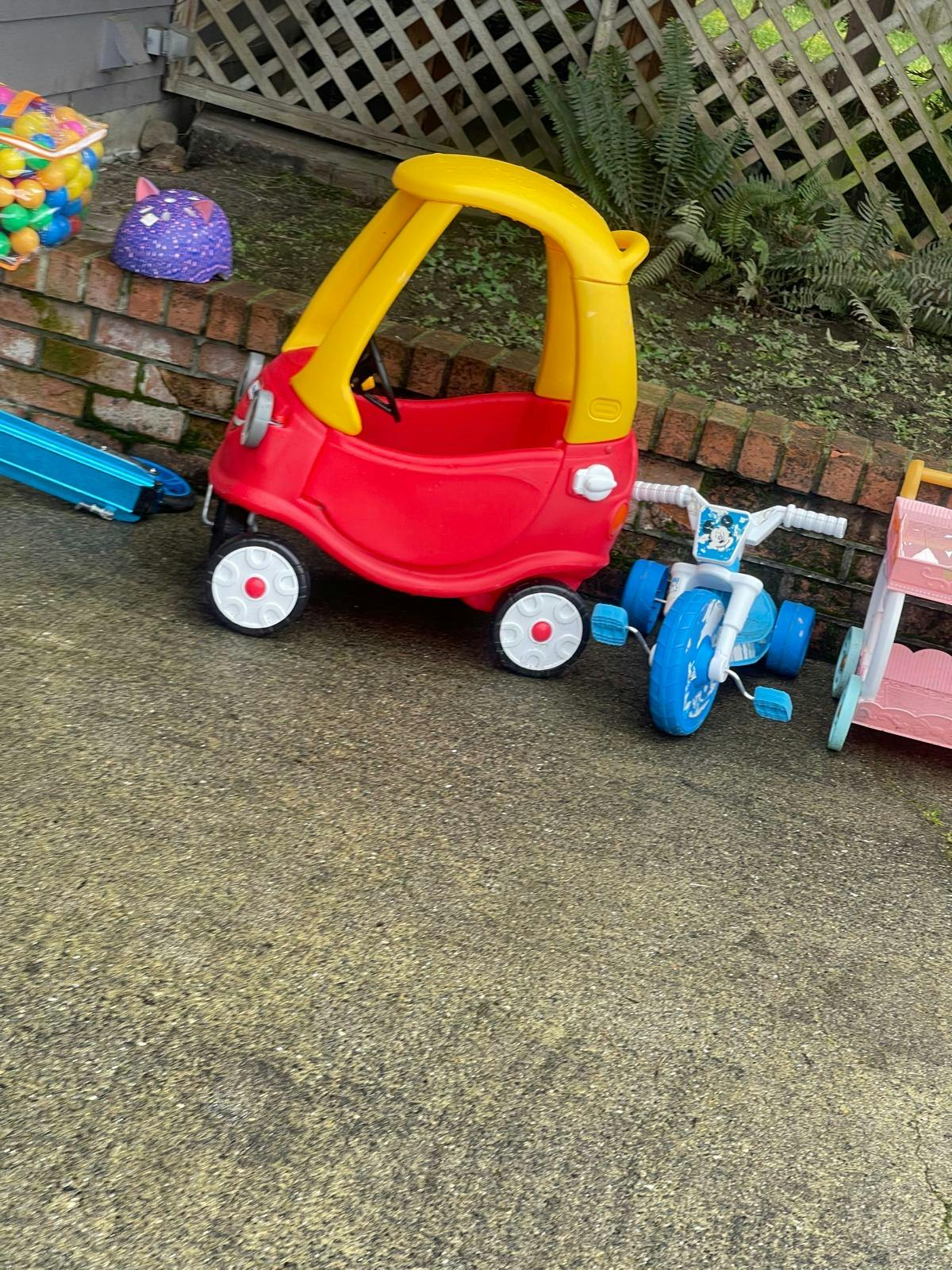 Red and yellow toy car next to a blue tricycle and pink toy cart