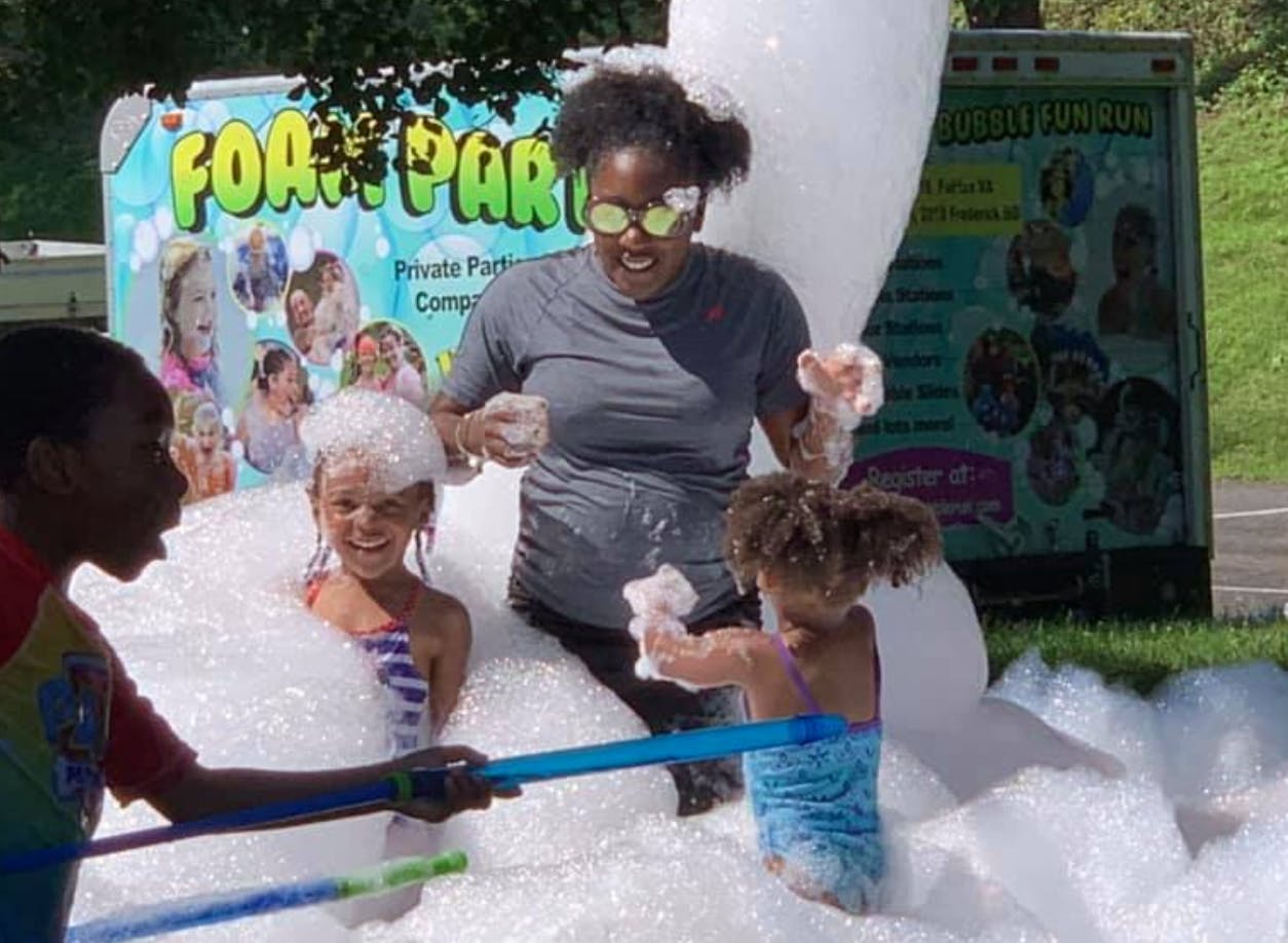 Adult and children playing in a foam party with a "Foam Party" sign in the background
