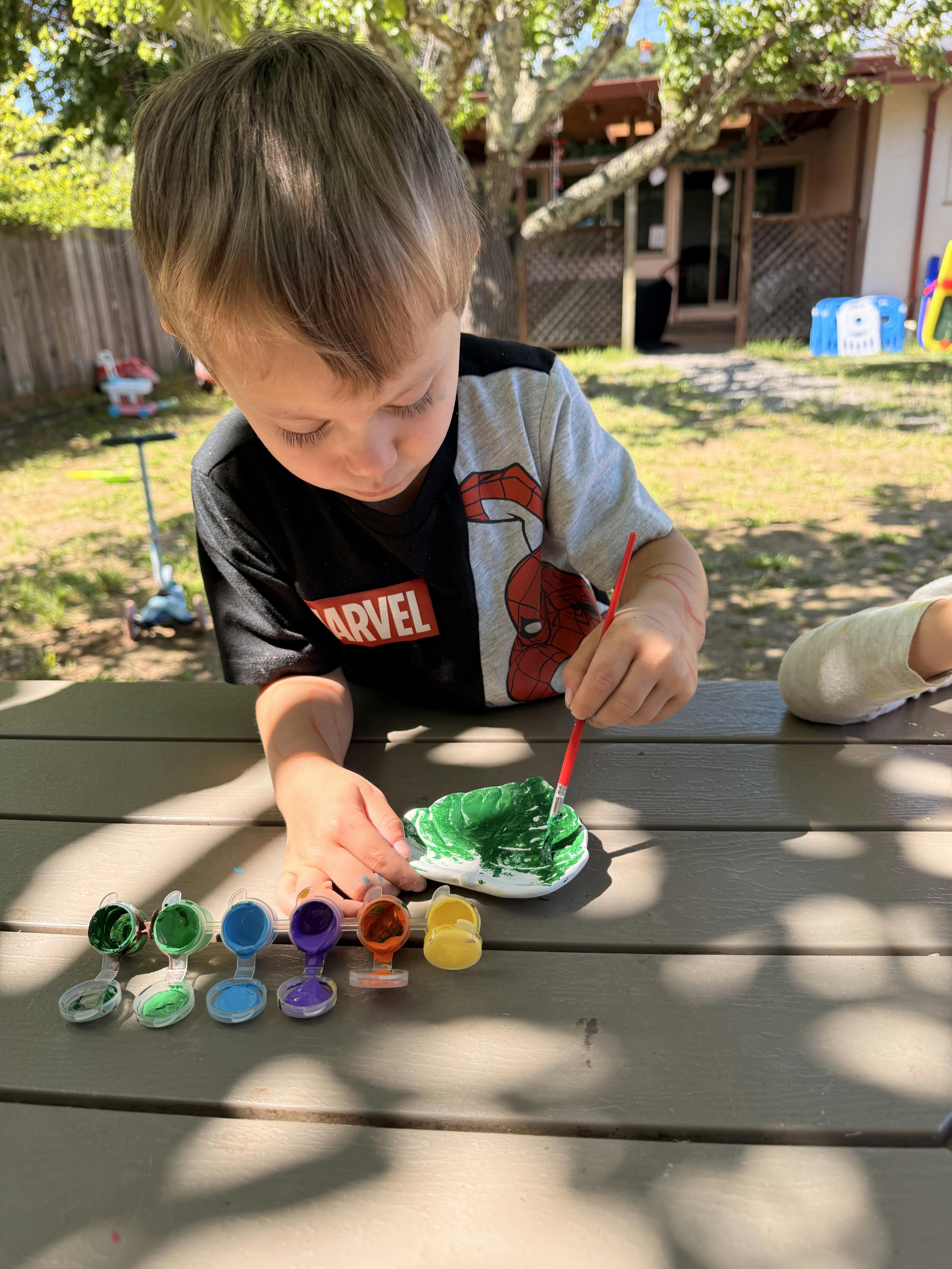 Child painting a white object green at a table outdoors