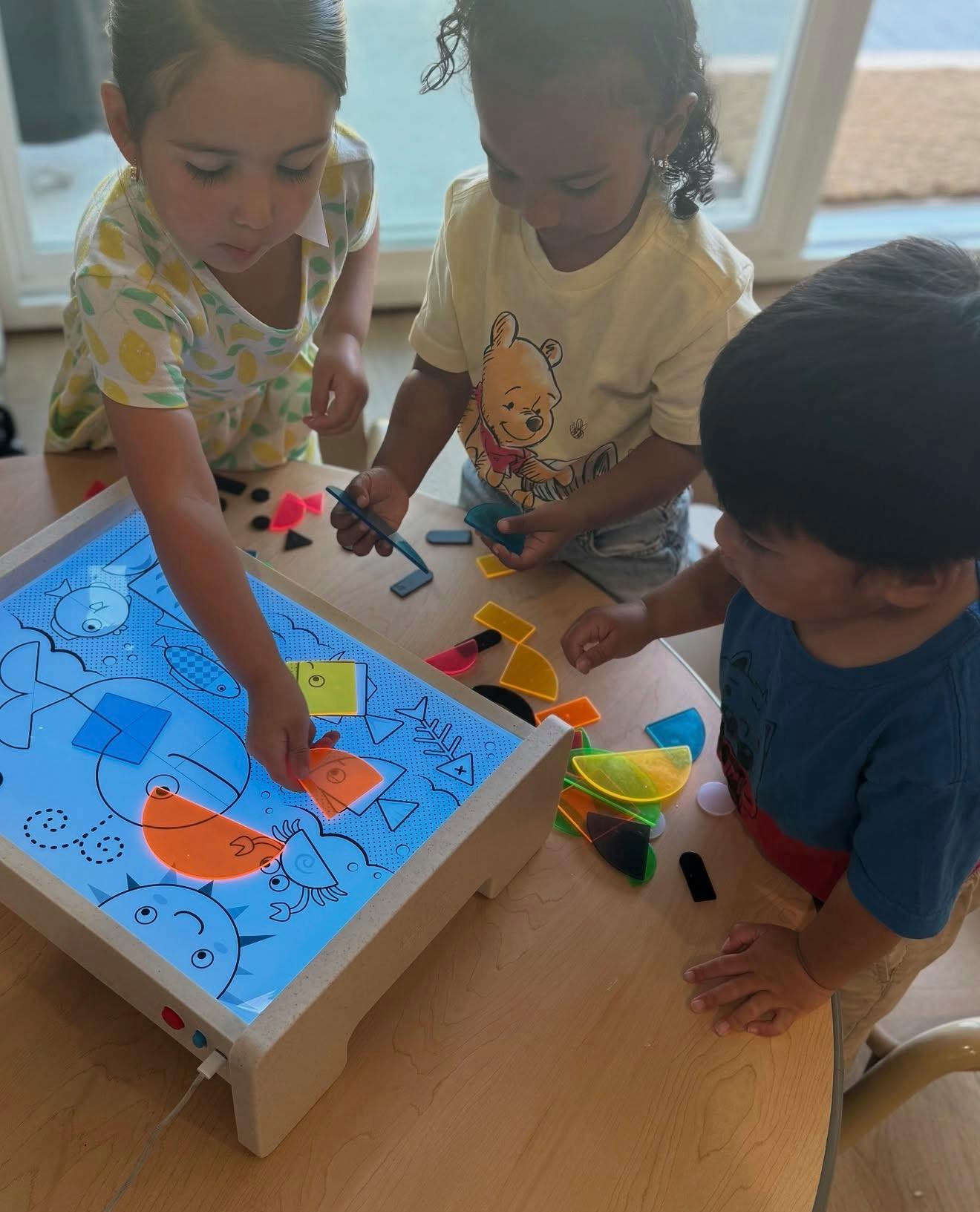Three children playing with colorful translucent shapes on a light table