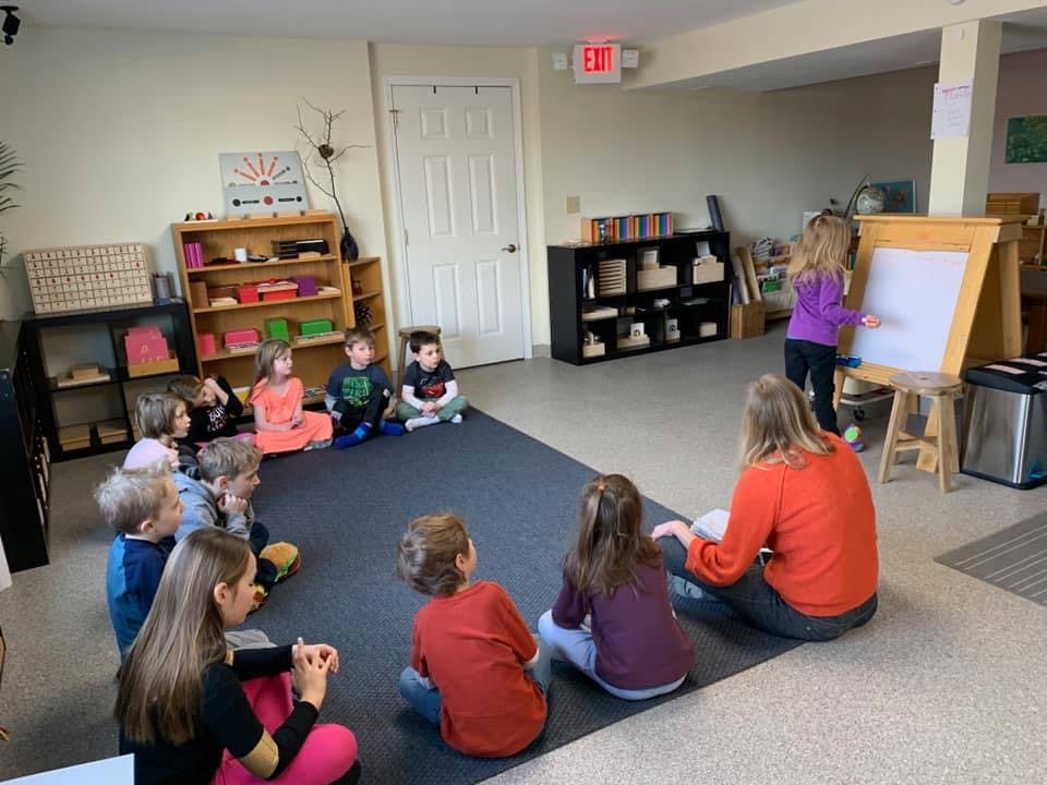 Adults and children sitting on a rug in a classroom, one child at an easel