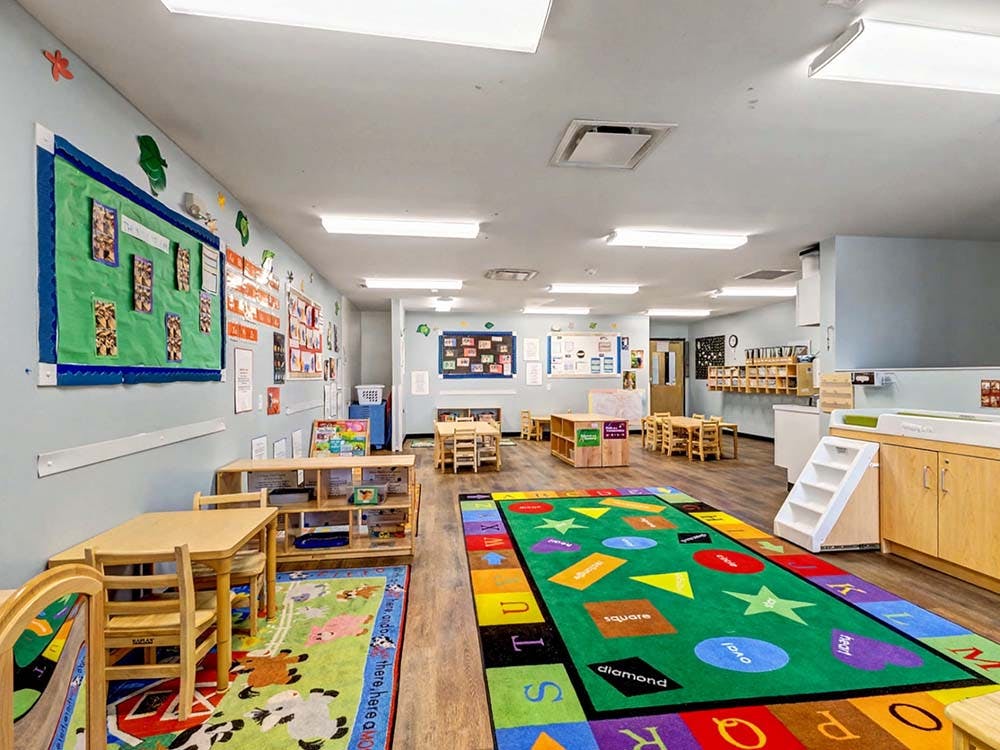 Classroom with small tables and chairs, wall art, and a colorful rug with shapes