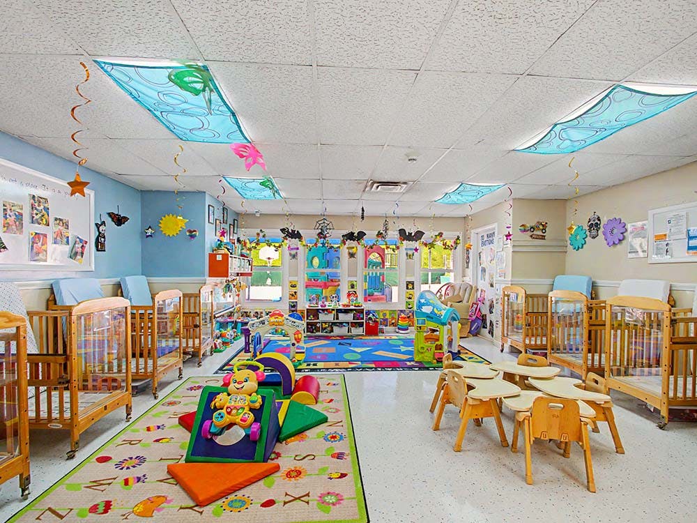 A colorful childcare classroom with cribs, play mats, and small tables